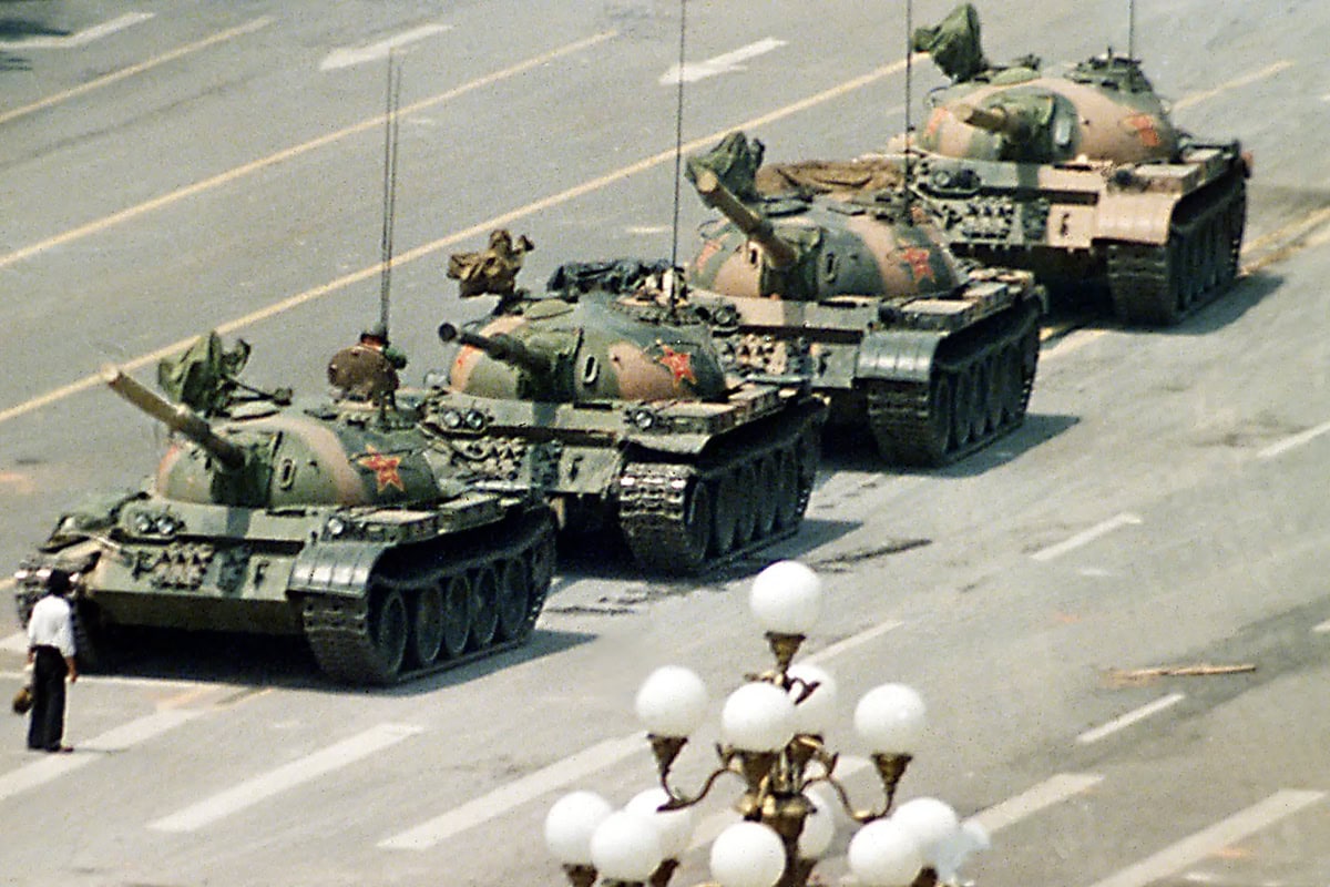 Color photograph of a man standing in front of four military tanks on an empty street.