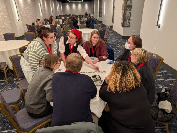 A group of eight people gathered around a roundtable playing a card game