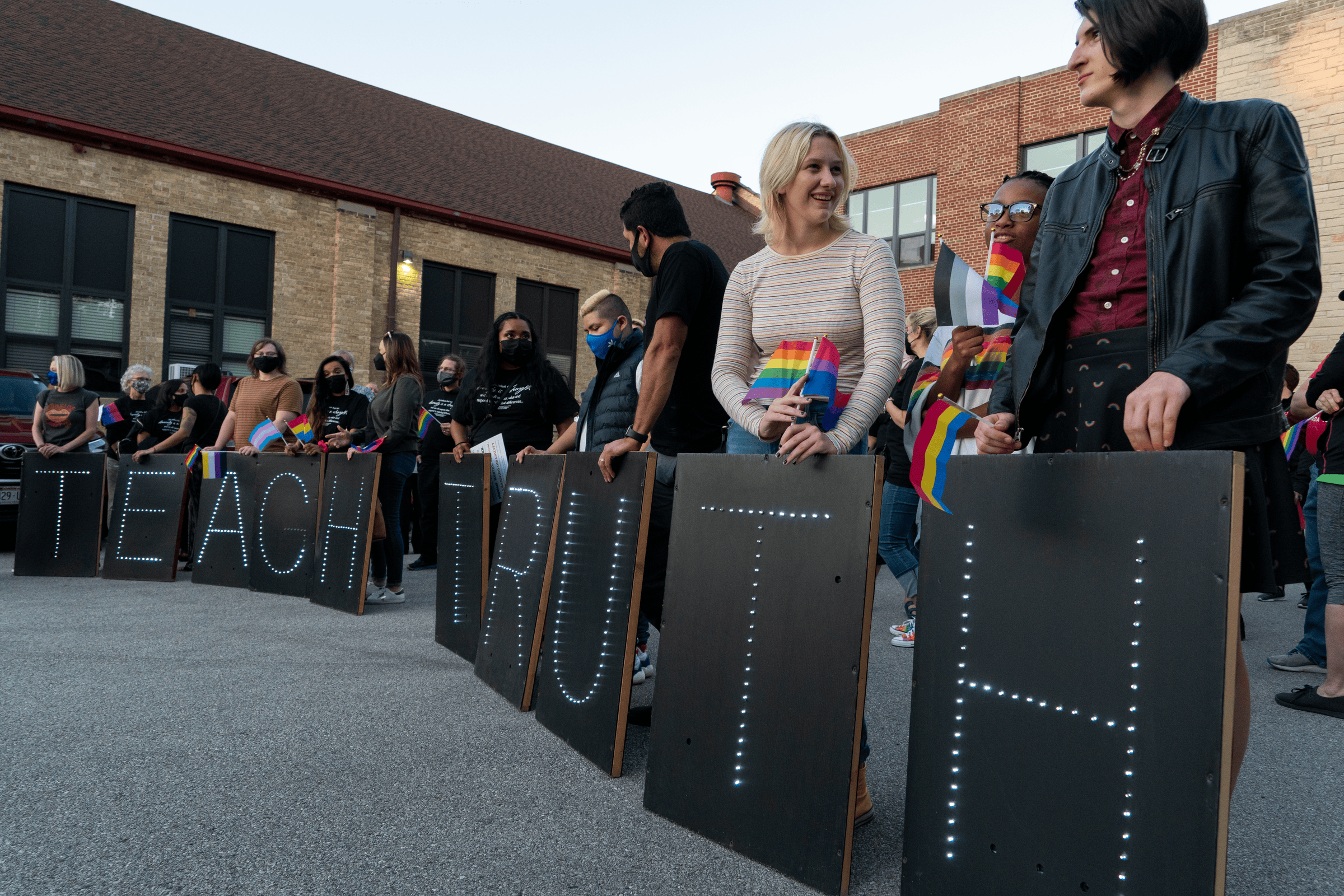 A group of people standing in a line wearing rainbow masks and holding signs in front of them that spell out 'Teach Truth'